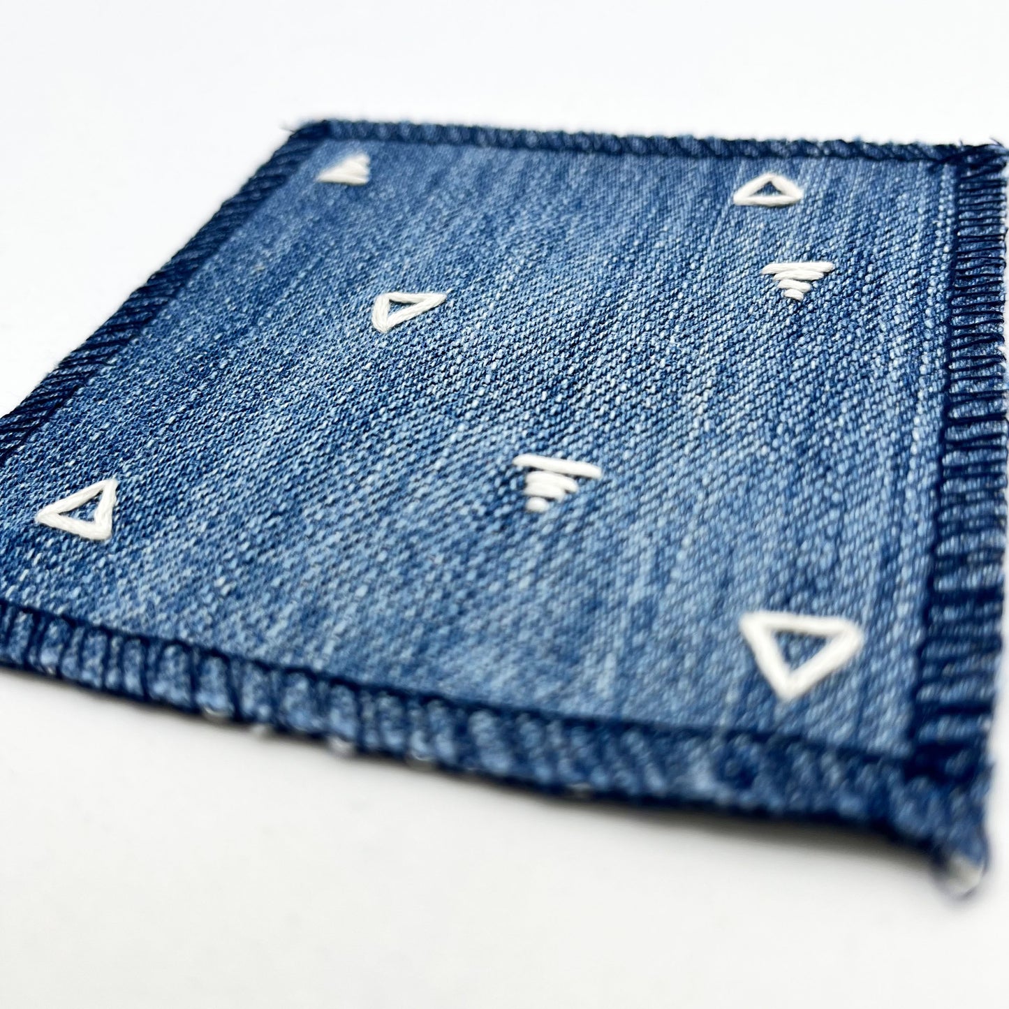 an angled close up view of a square patch made out of denim, handstitched with scattered ivory triangles, some as outlines some with a line fill, with overlocked edges, on a white background