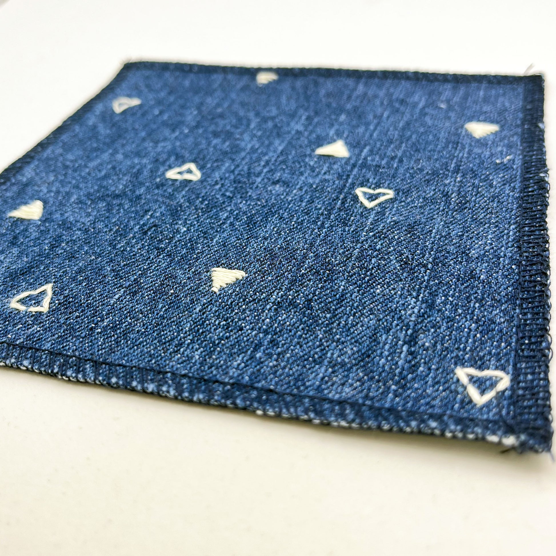 an angled close up view of a square patch made out of denim, handstitched with scattered ivory triangles, some as outlines some with a line fill, with overlocked edges, on a white background