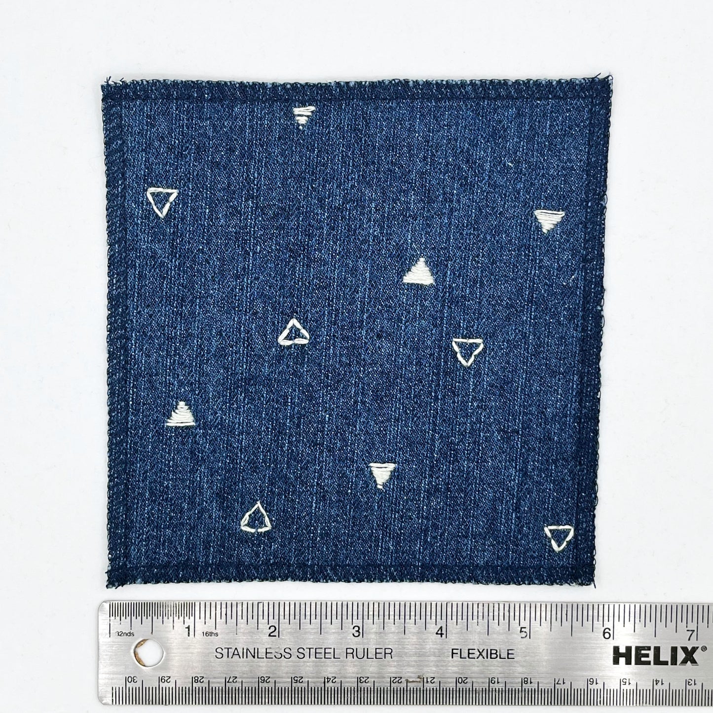 a square patch made out of denim, handstitched with scattered ivory triangles, some as outlines some with a line fill, with overlocked edges, on a white background, next to a ruler showing a width of 6 inches