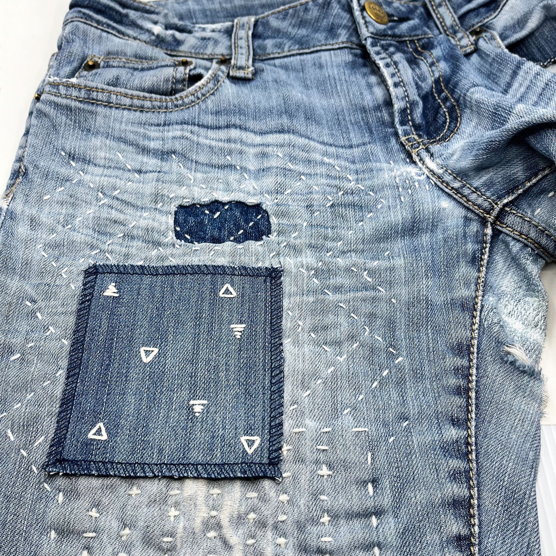 a square patch made out of denim, handstitched with scattered ivory triangles, some as outlines some with a line fill, with overlocked edges, on jeans with other visible mending and sashiko stitching