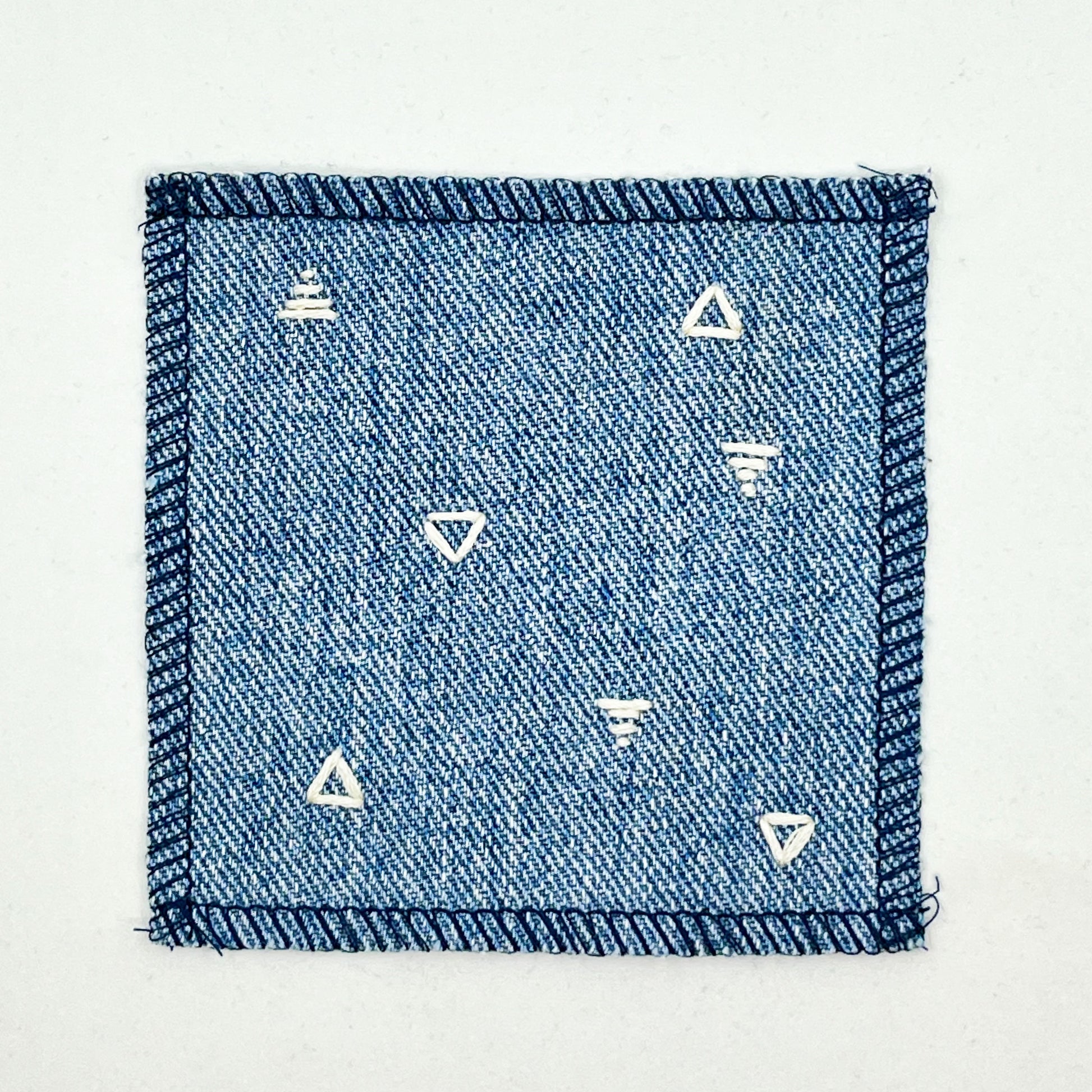 a square patch made out of denim, handstitched with scattered ivory triangles, some as outlines some with a line fill, with overlocked edges, on a white background