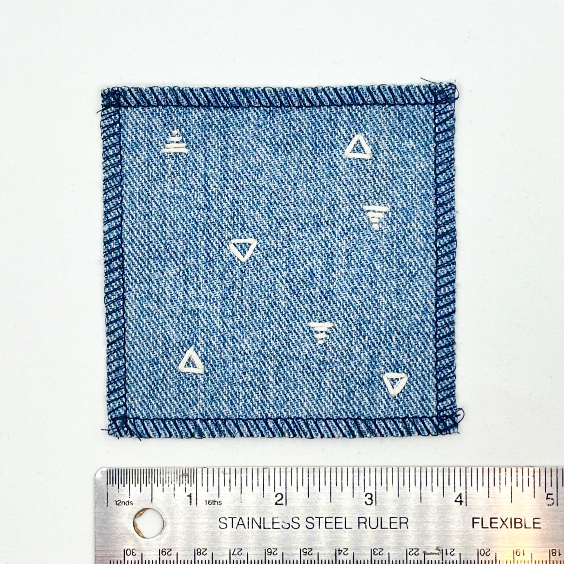 a square patch made out of denim, handstitched with scattered ivory triangles, some as outlines some with a line fill, with overlocked edges, on a white background, next to a ruler showing a width of 4 inches
