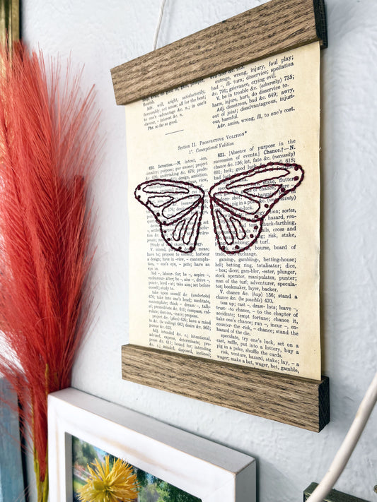a vintage thesaurus page in a magnetic wood frame hanging on a wall next to other framed photos and decorative branches and feathers, the book page is hand embroidered in plum colored thread with butterfly wings