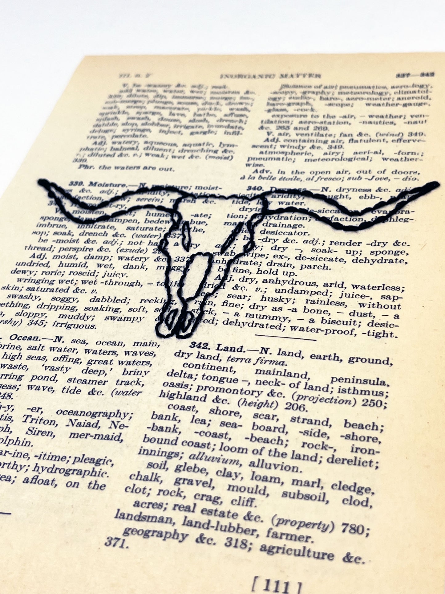 angled view of a vintage thesaurus page hand embroidered in black thread with a longhorn skull on a white background