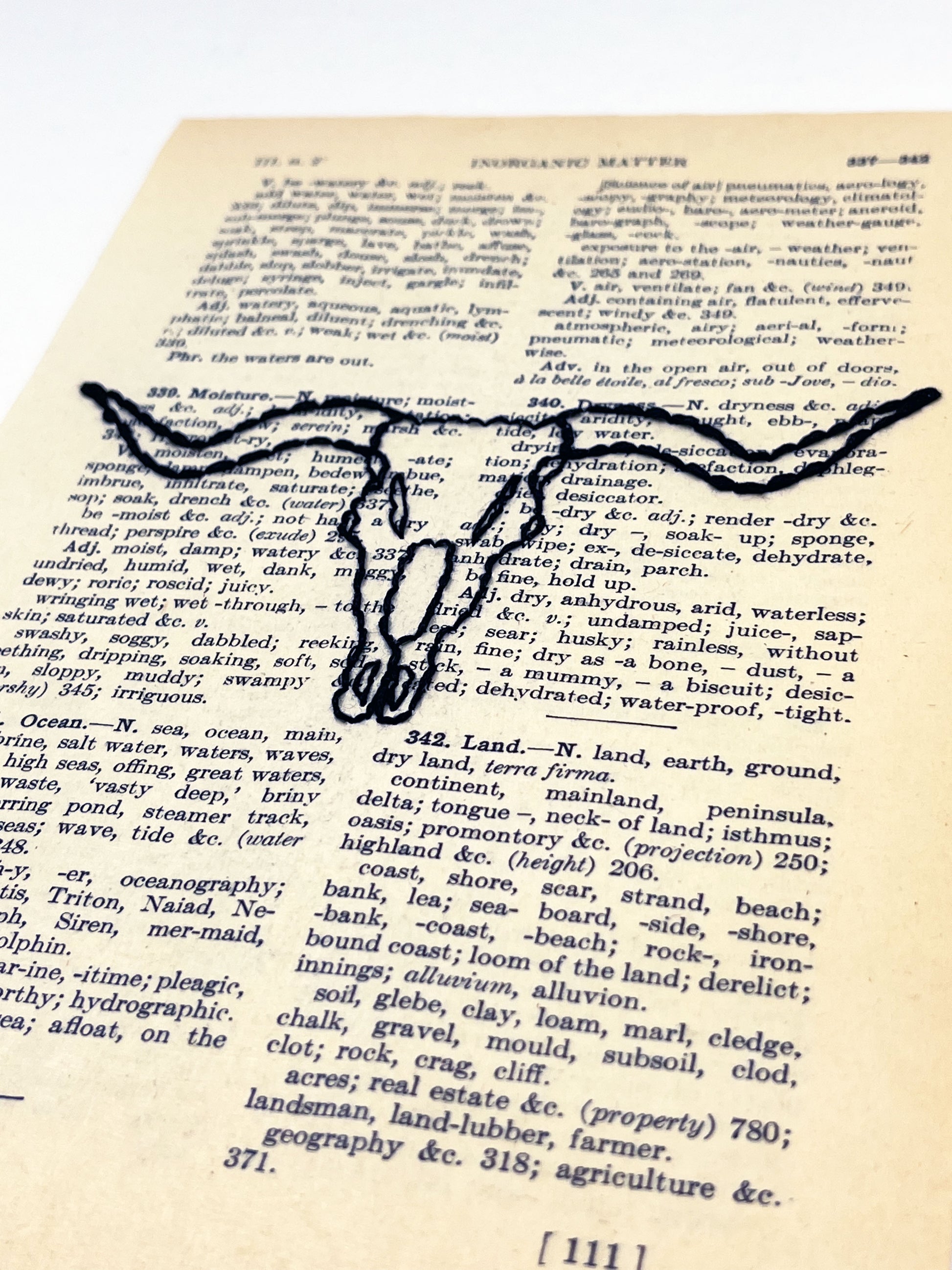angled view of a vintage thesaurus page hand embroidered in black thread with a longhorn skull on a white background