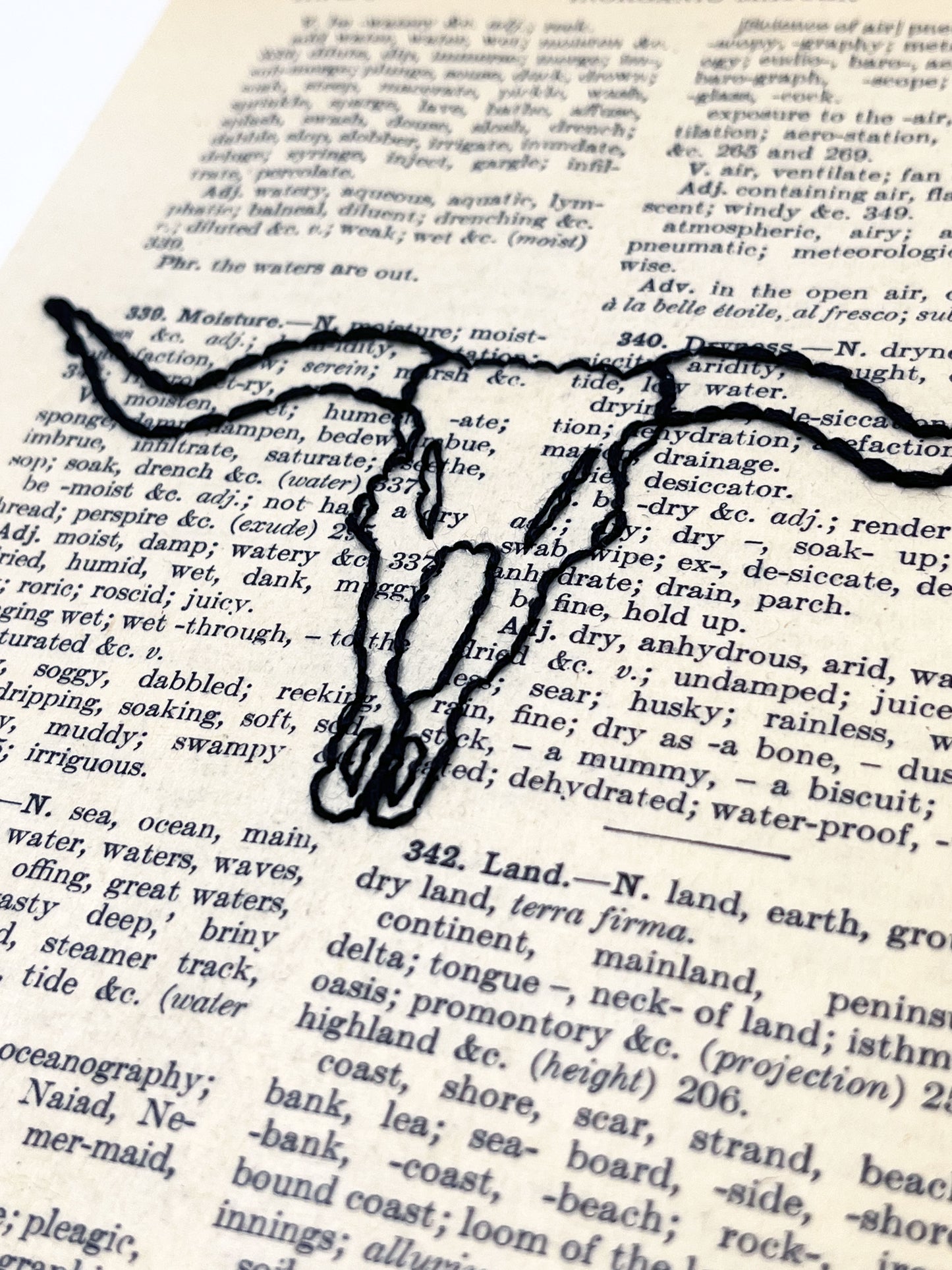 close up view of a vintage thesaurus page hand embroidered in black thread with a longhorn skull on a white background