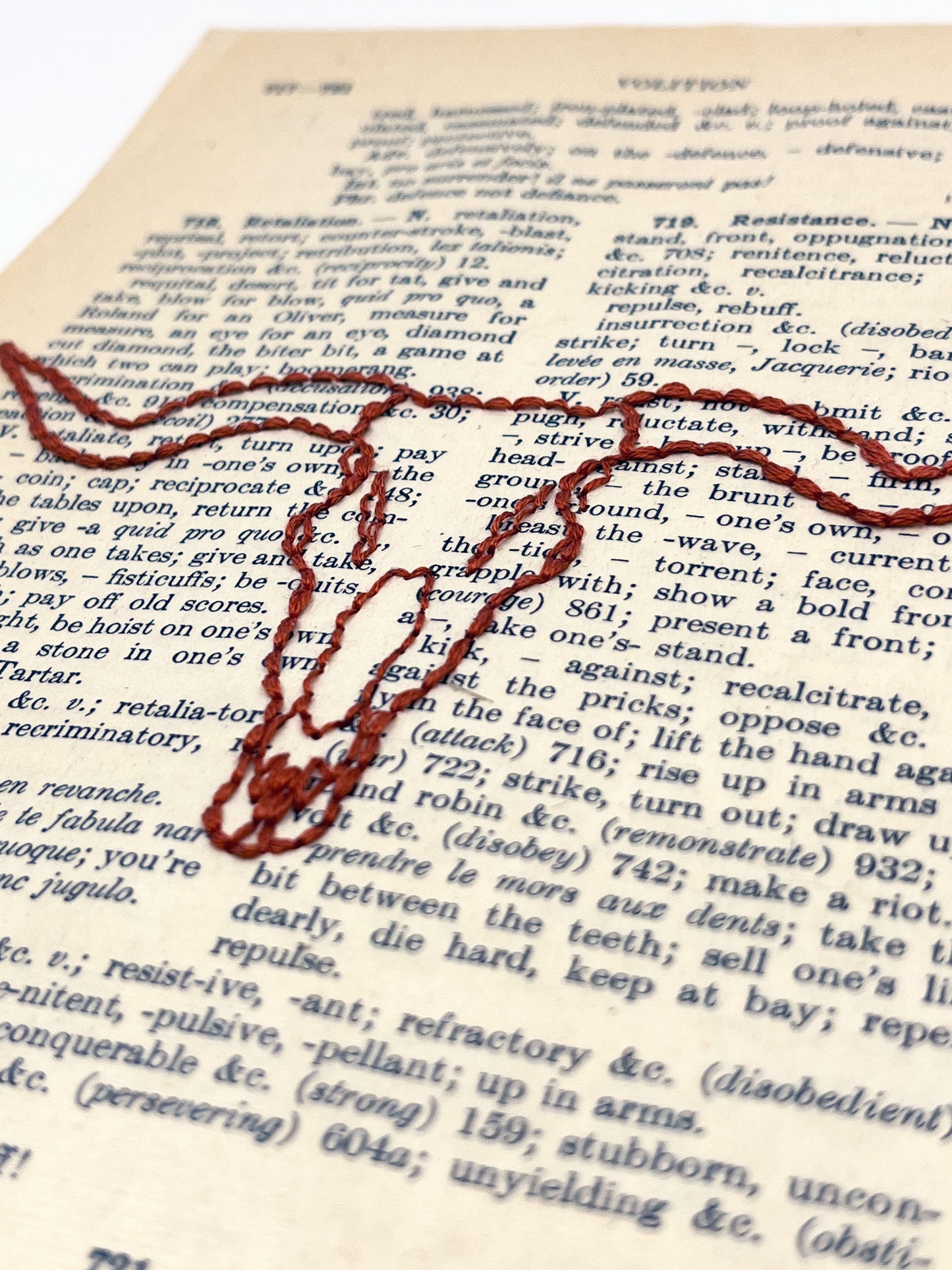 close up view of a vintage thesaurus page hand embroidered in rust colored thread with a longhorn skull on a white background