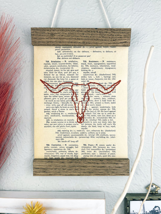 a vintage thesaurus page in a magnetic wood frame hanging on a wall next to other framed photos and decorative branches and feathers, the book page is hand embroidered in rust colored thread with a longhorn skull