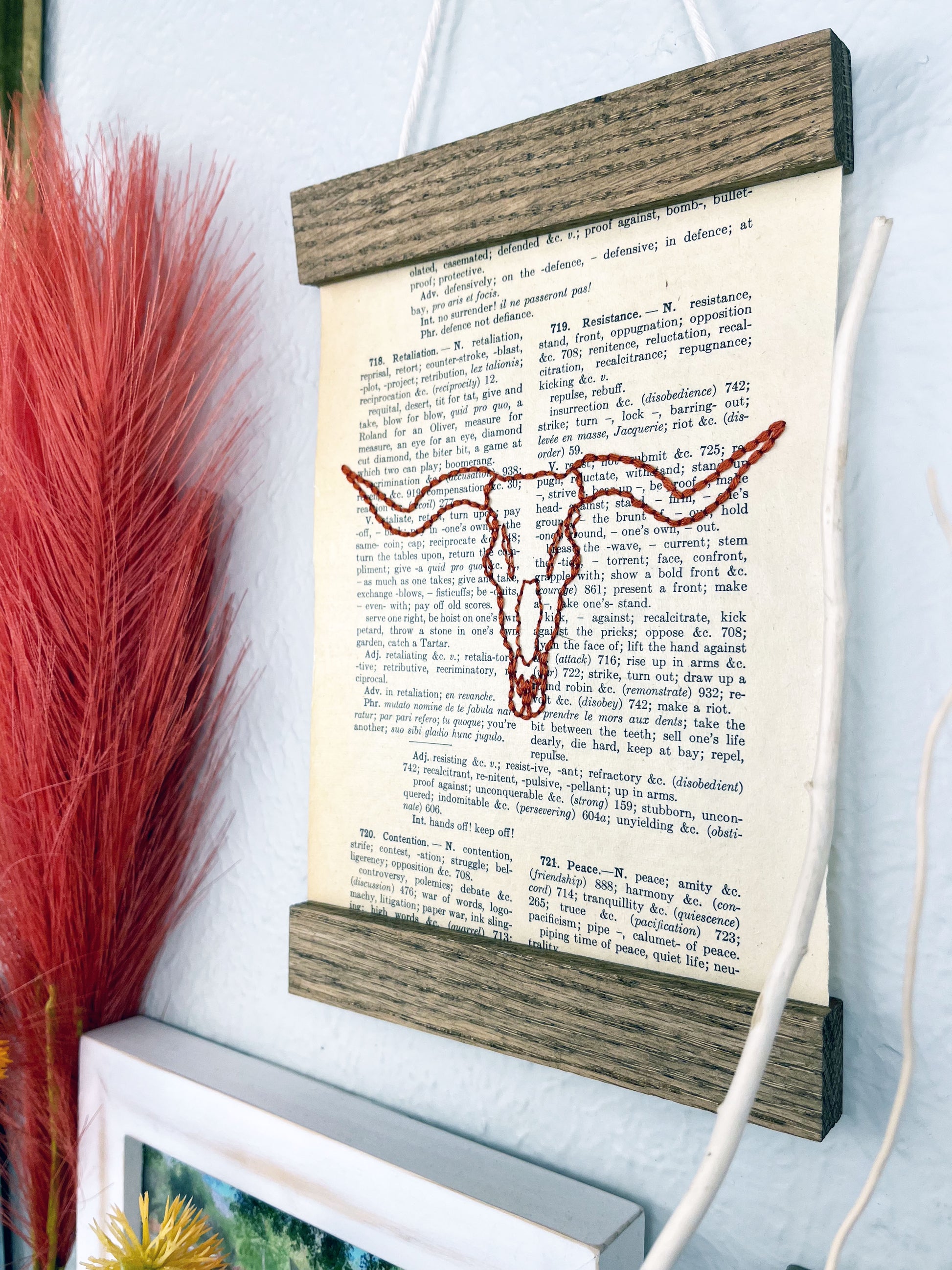 a vintage thesaurus page in a magnetic wood frame hanging on a wall next to other framed photos and decorative branches and feathers, the book page is hand embroidered in rust colored thread with a longhorn skull