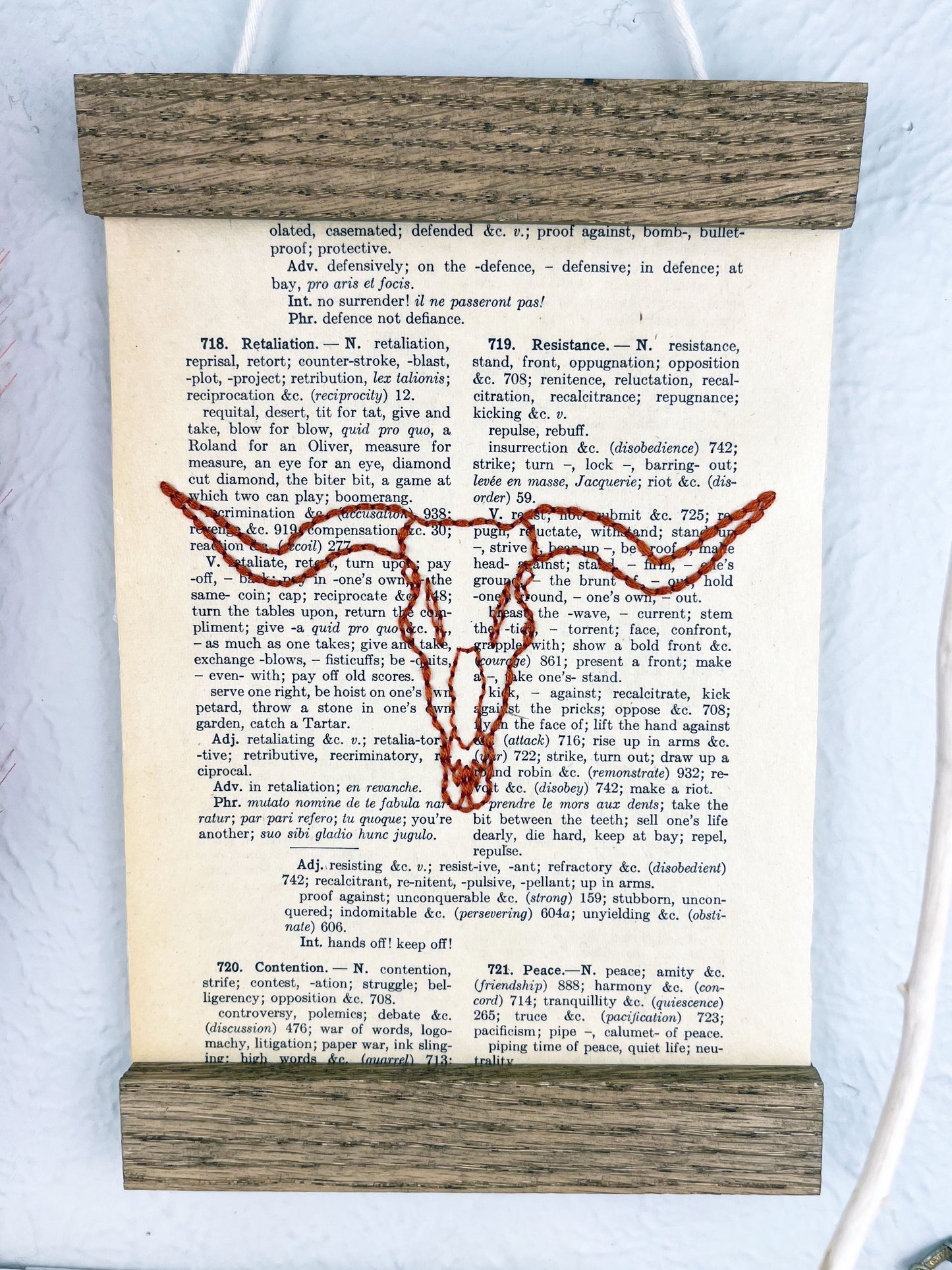 a vintage thesaurus page in a magnetic wood frame hanging on a wall hand embroidered in rust colored thread with a longhorn skull