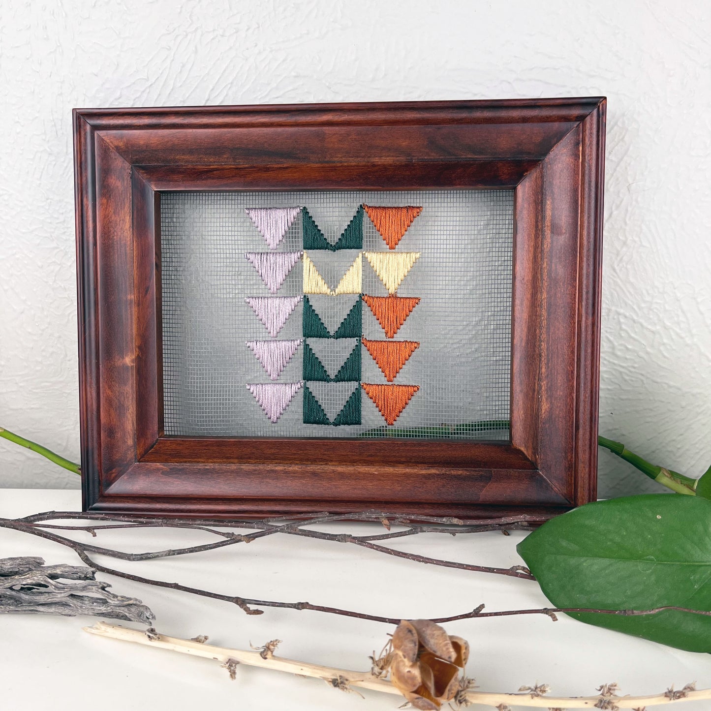 a piece of window screen hand stitched with 3 rows of triangles resembling the flying geese quilt blocks, in light purple pine green butter yellow and rust brown, in a wood frame, on a white counter, surrounded by a pothos vine, dried flowers and sticks