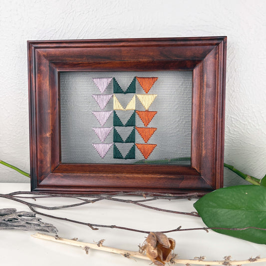 a piece of window screen hand stitched with 3 rows of triangles resembling the flying geese quilt blocks, in light purple pine green butter yellow and rust brown, in a wood frame, on a white counter, surrounded by a pothos vine, dried flowers and sticks