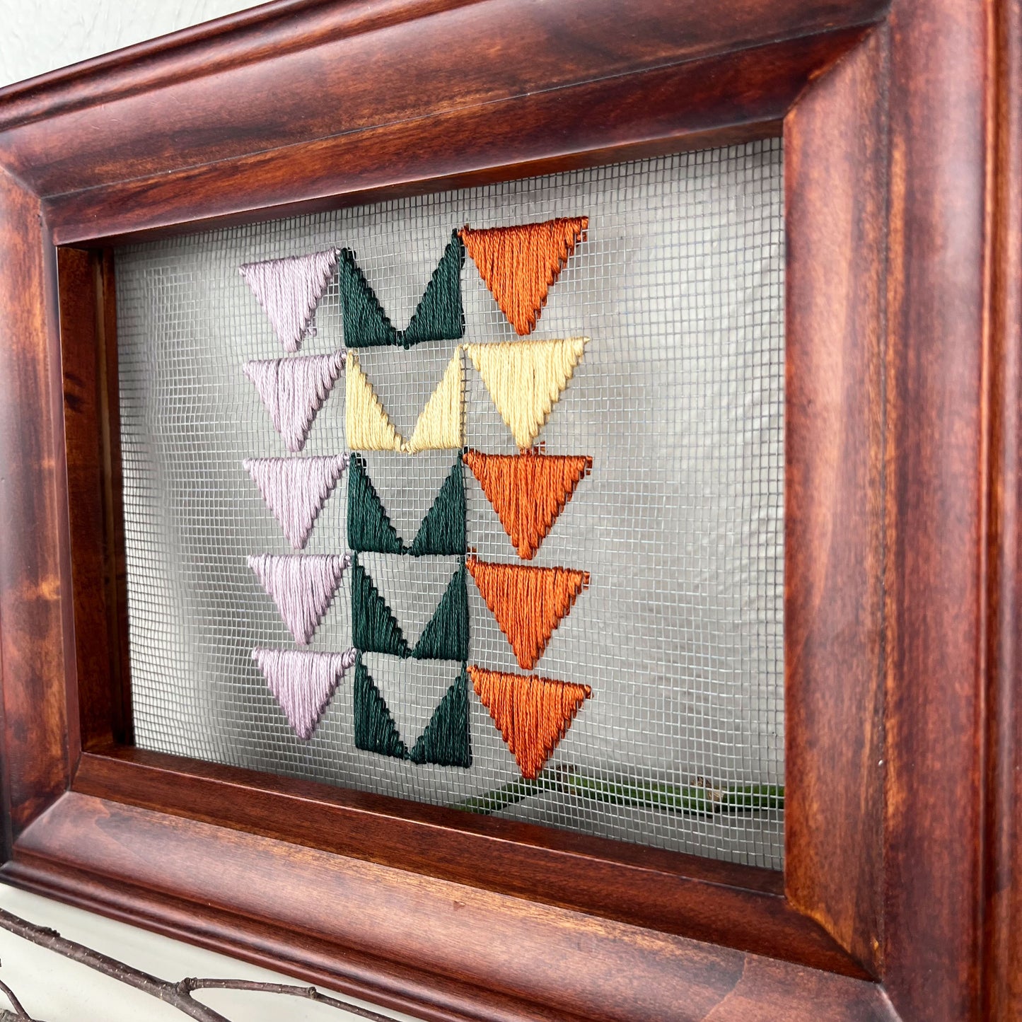 close up angled view of a piece of window screen hand stitched with 3 rows of triangles resembling the flying geese quilt blocks, in light purple pine green wheat yellow and rust brown, in a wood frame, on a white counter, surrounded by a pothos vine, dried flowers and sticks