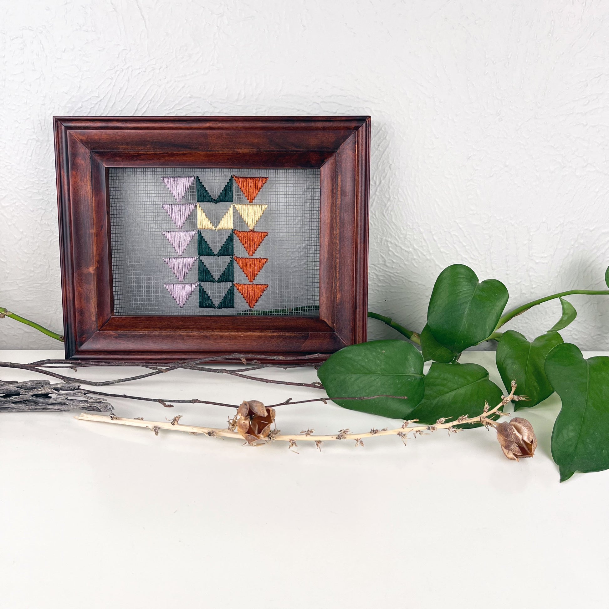 a piece of window screen hand stitched with 3 rows of triangles resembling the flying geese quilt blocks, in light purple pine green wheat yellow and rust brown, in a wood frame, on a white counter, surrounded by a pothos vine, dried flowers and sticks