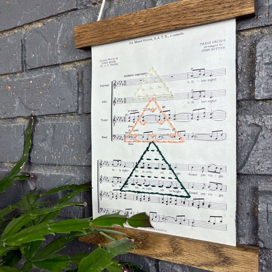 Christmas sheet music, hand stitched on with a Christmas tree made from triangles filled with different types of stitches, in green, peach and ivory thread, hanging in a wood magnetic frame on a grey brick wall, with a Christmas cactus in front of it