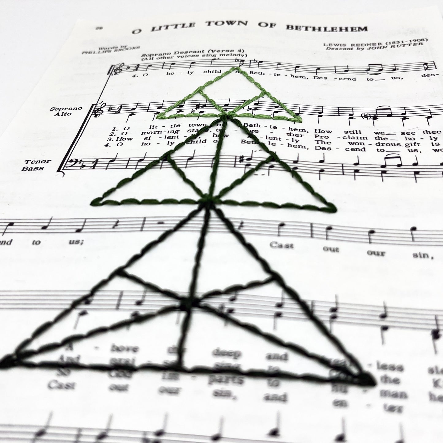 a close up angled view of sheet music from the Christmas "O Little Town of Bethlehem", hand stitched over with a Christmas tree made from triangles, in shades of green thread