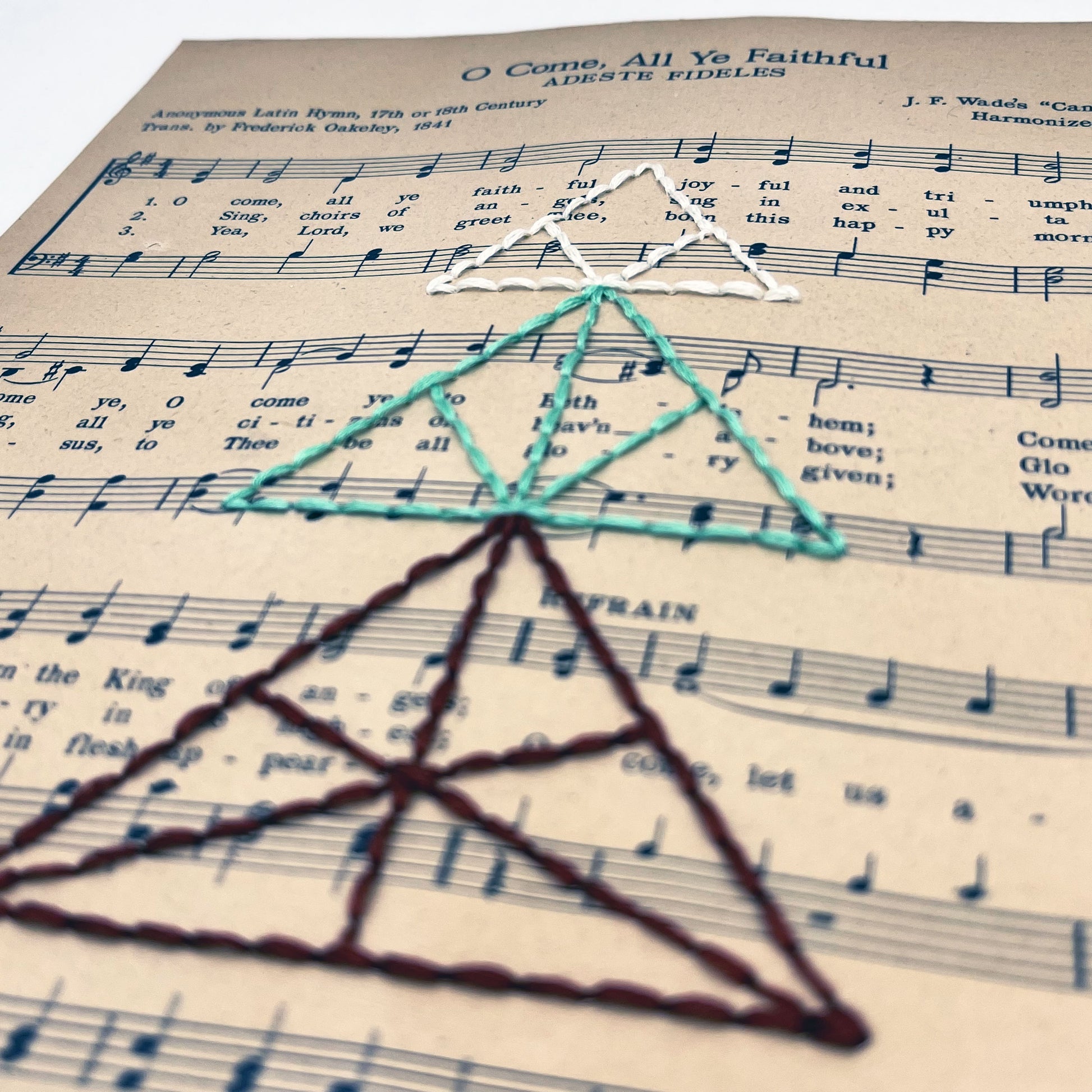 a close up angled view of sheet music from the Christmas song O Come All Ye Faithful, hand stitched over with a Christmas tree made from triangles, in red, green and ivory thread