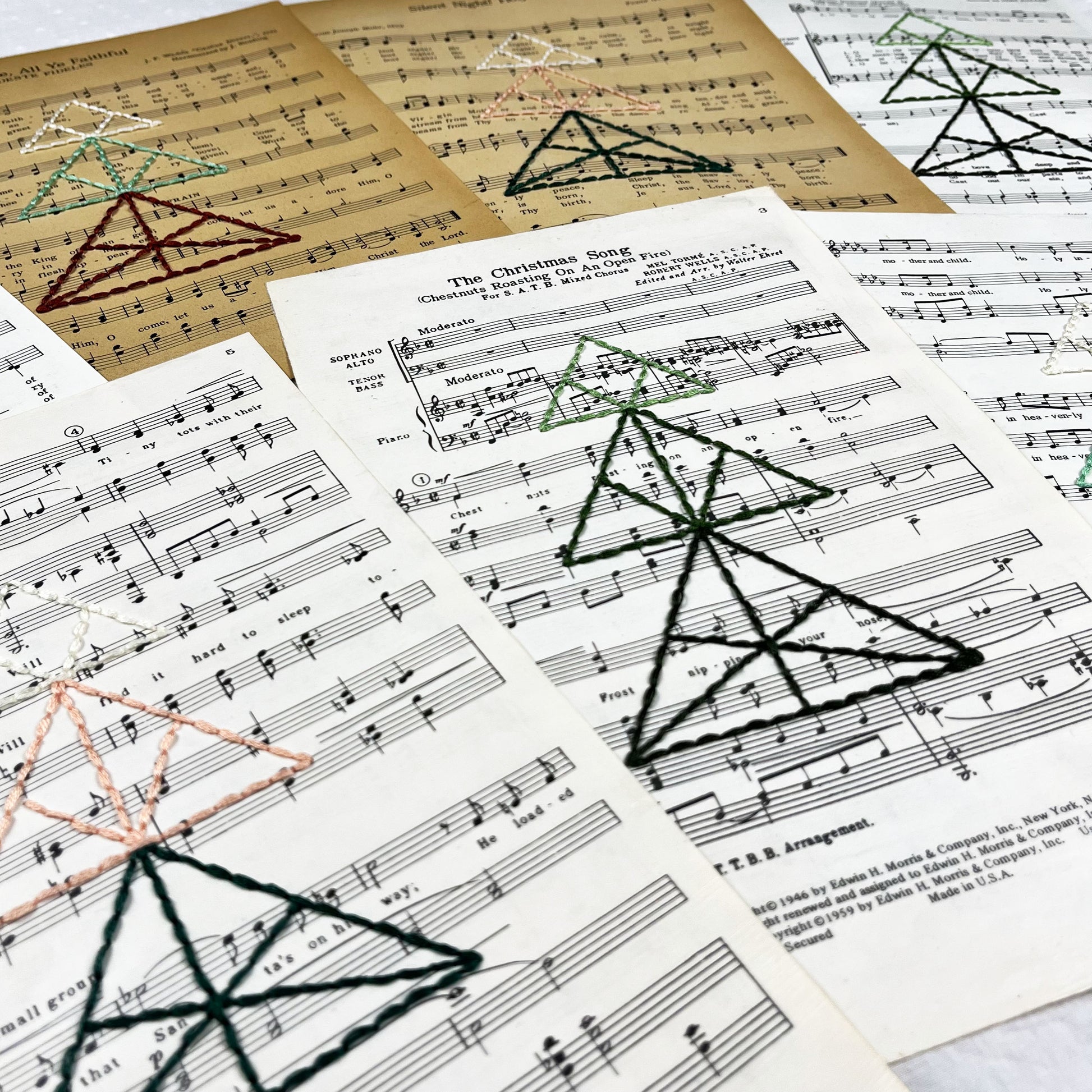 close up angled view of a group of sheet music from Christmas songs, hand stitched over with a Christmas tree made from triangles, in different color combinations
