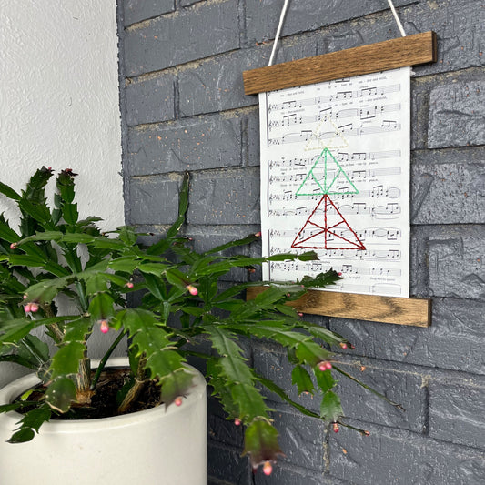 sheet music from a Christmas song, hand stitched over with a Christmas tree made from triangles, in red green and ivory thread, hanging in a wood magnetic frame on a grey brick wall, with a Christmas cactus in the foreground