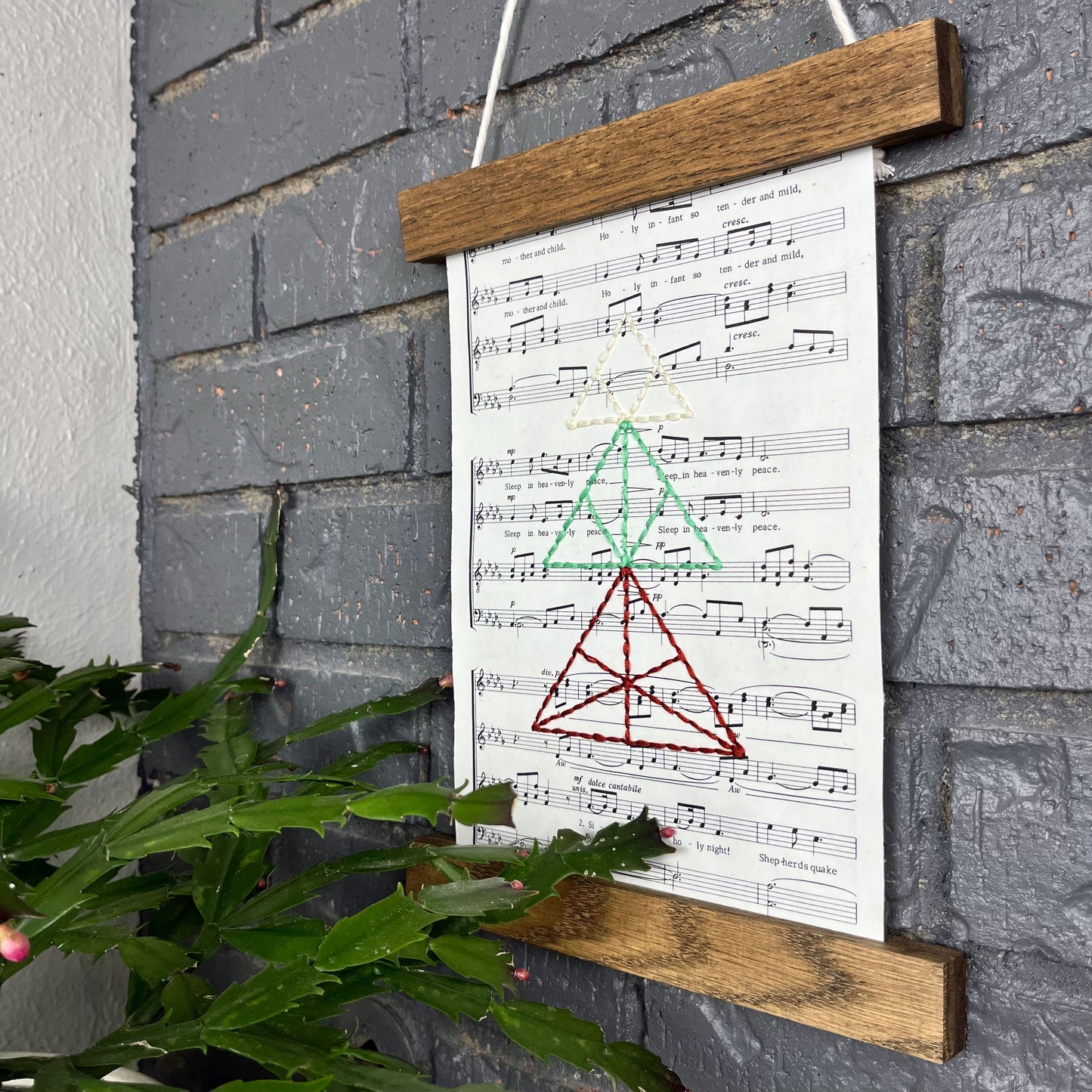 sheet music from a Christmas song, hand stitched over with a Christmas tree made from triangles, in red green and ivory thread, hanging in a wood magnetic frame on a grey brick wall, with a Christmas cactus in the foreground