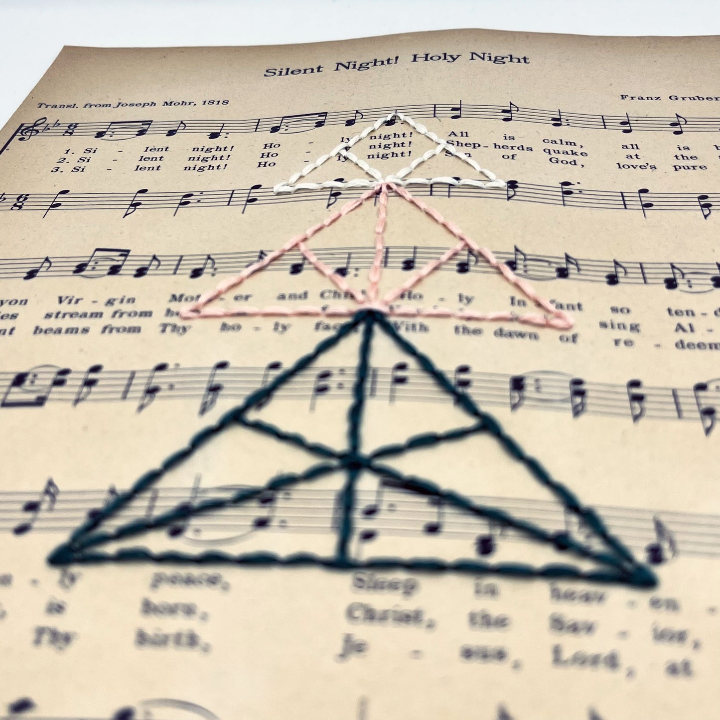a close up angled view of sheet music from the Christmas song "Silent Night", hand stitched over with a Christmas tree made from triangles, in green, peach and ivory thread