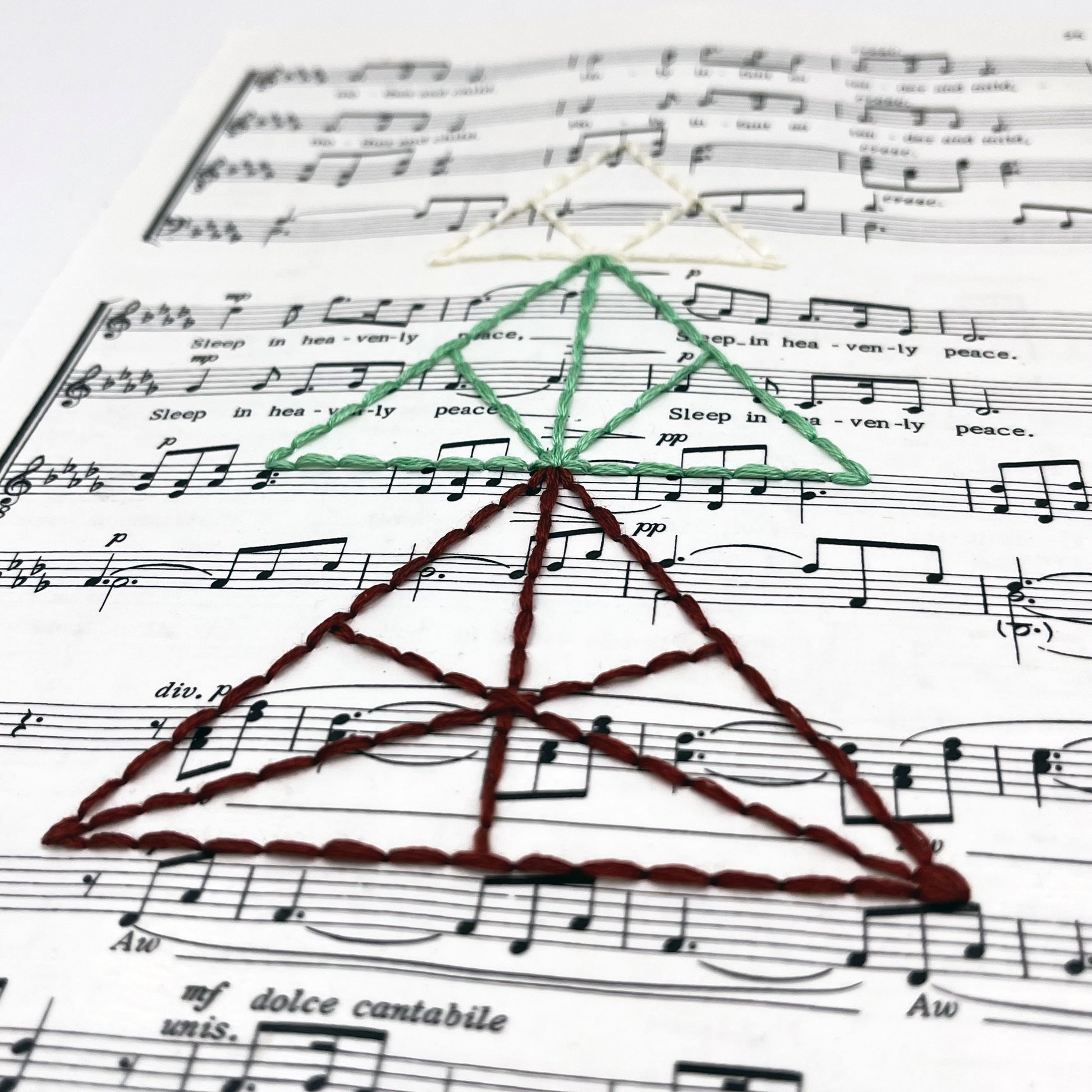 a close up angled view of sheet music from the Christmas song "Silent Night", hand stitched over with a Christmas tree made from triangles, in red, green and ivory thread
