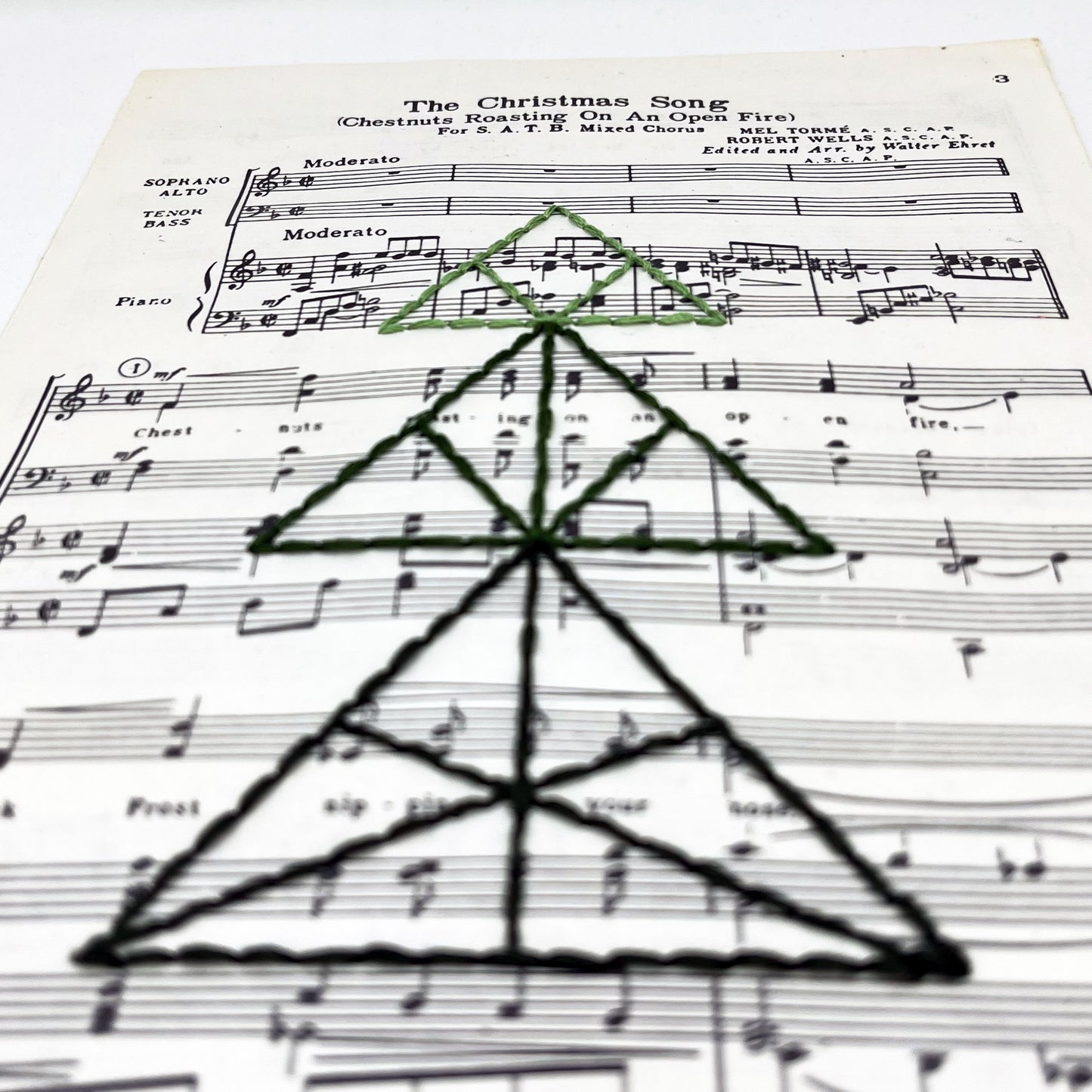 a close up angled view of sheet music from "The Christmas Song", hand stitched over with a Christmas tree made from triangles, in shades of green thread