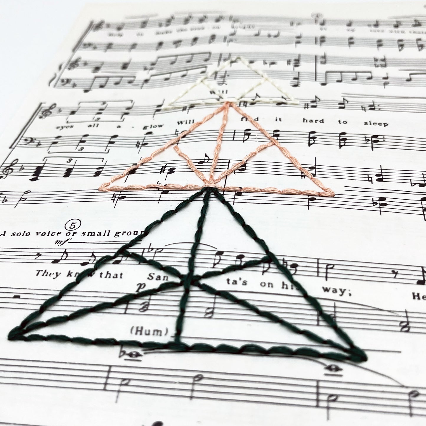 a close up angled view of sheet music from "The Christmas Song", hand stitched over with a Christmas tree made from triangles, in green, peach and ivory thread