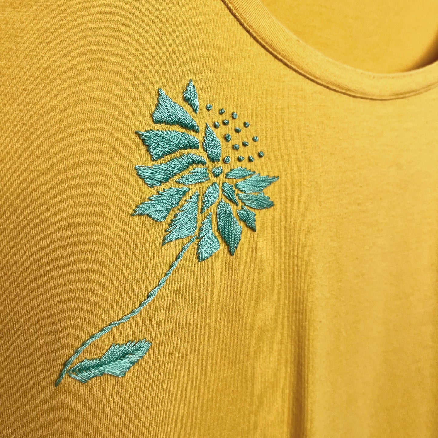 a hand embroidered abstract aqua colored dandelion on a mustard yellow t-shirt, close up
