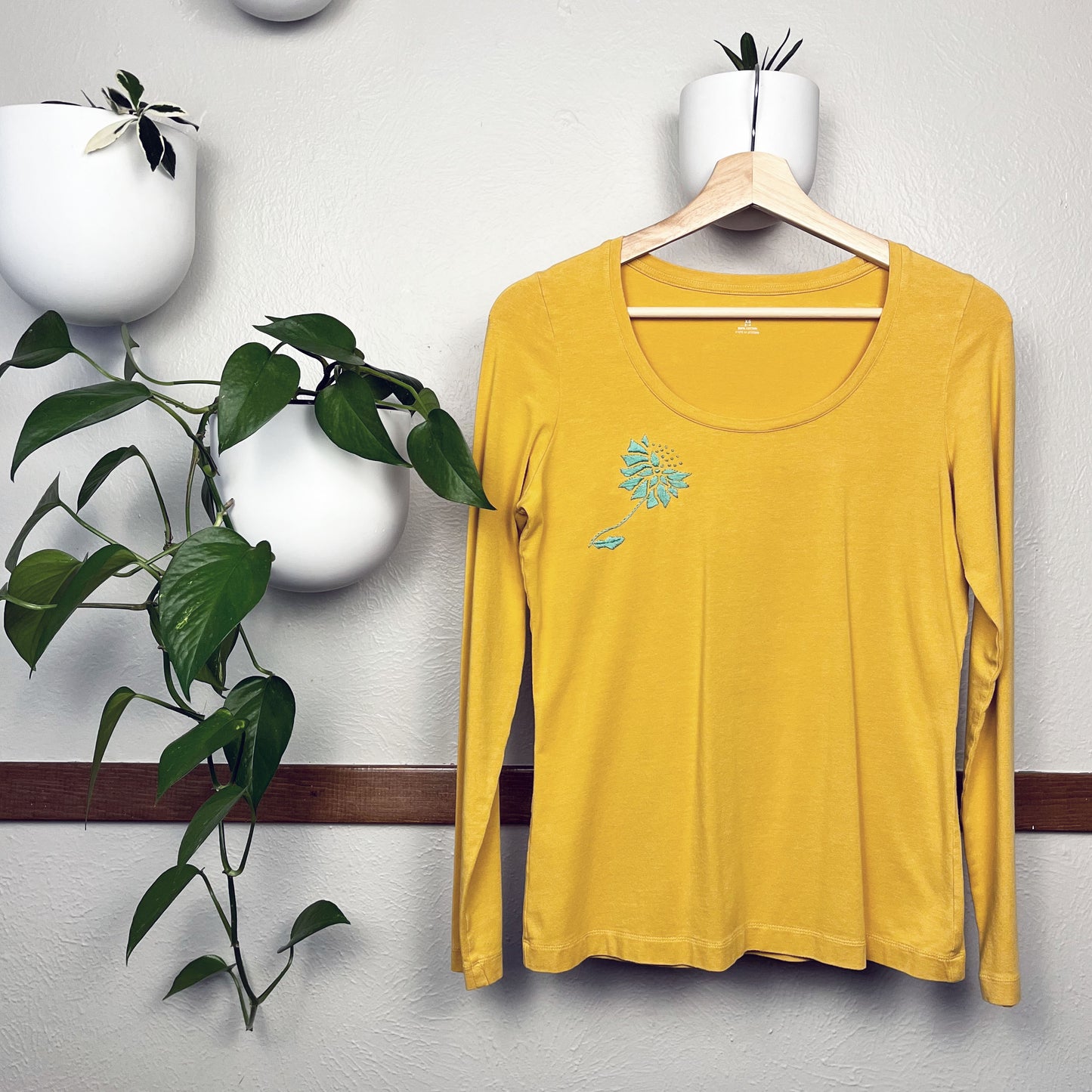 a bright mustard colored long sleeve tee, with a hand embroidered abstract aqua colored dandelion. hanging on a wooden hanger, with plants in white round bottomed pots hanging on the wall around it