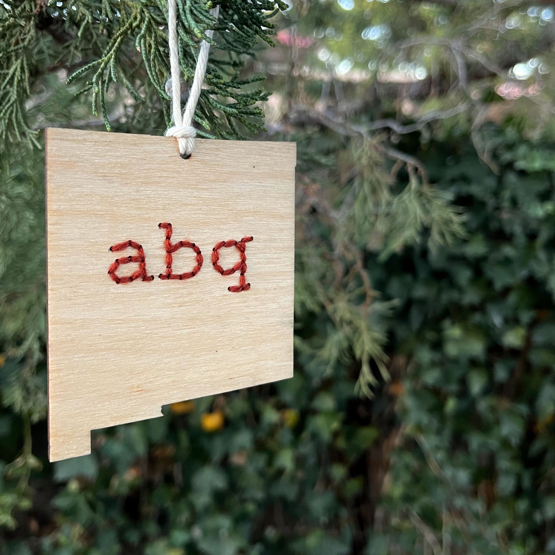 a laser cut wood ornament in the shape of New Mexico, hand embroidered in rust red with the letters abq for Albuquerque, with an off white cord loop hanging on a pine tree