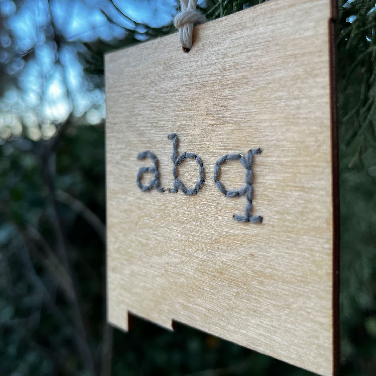 close up view of a laser cut wood ornament in the shape of New Mexico, hand embroidered in grey with the letters abq for Albuquerque, with an off white cord loop hanging on a pine tree
