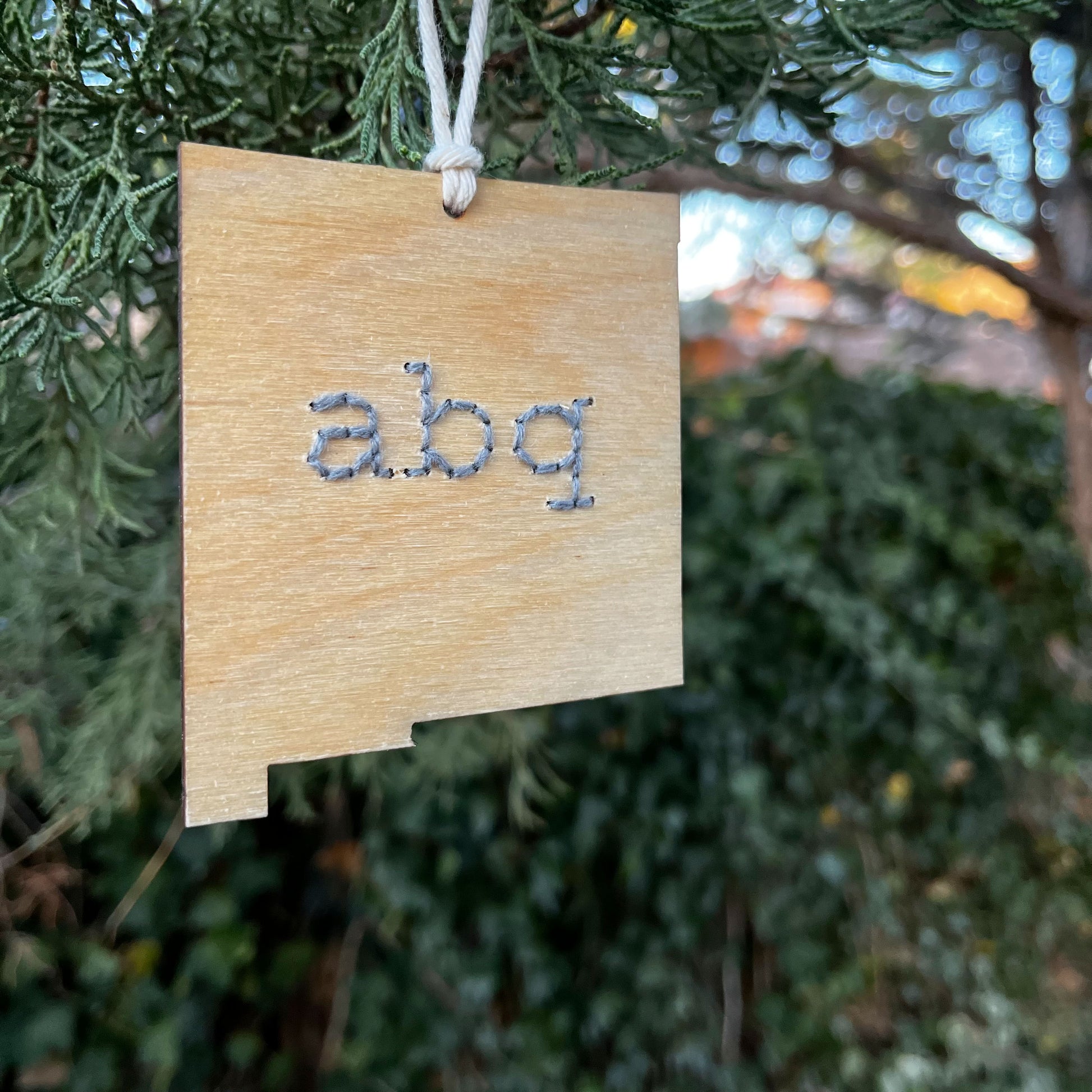 a laser cut wood ornament in the shape of New Mexico, hand embroidered in grey with the letters abq for Albuquerque, with an off white cord loop hanging on a pine tree