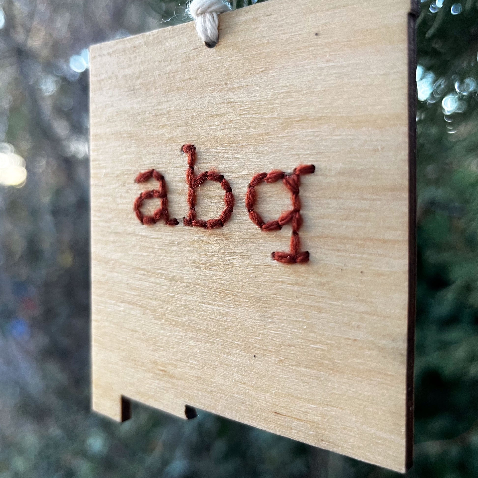 close up view of a laser cut wood ornament in the shape of New Mexico, hand embroidered in rust red with the letters abq for Albuquerque, with an off white cord loop hanging on a pine tree