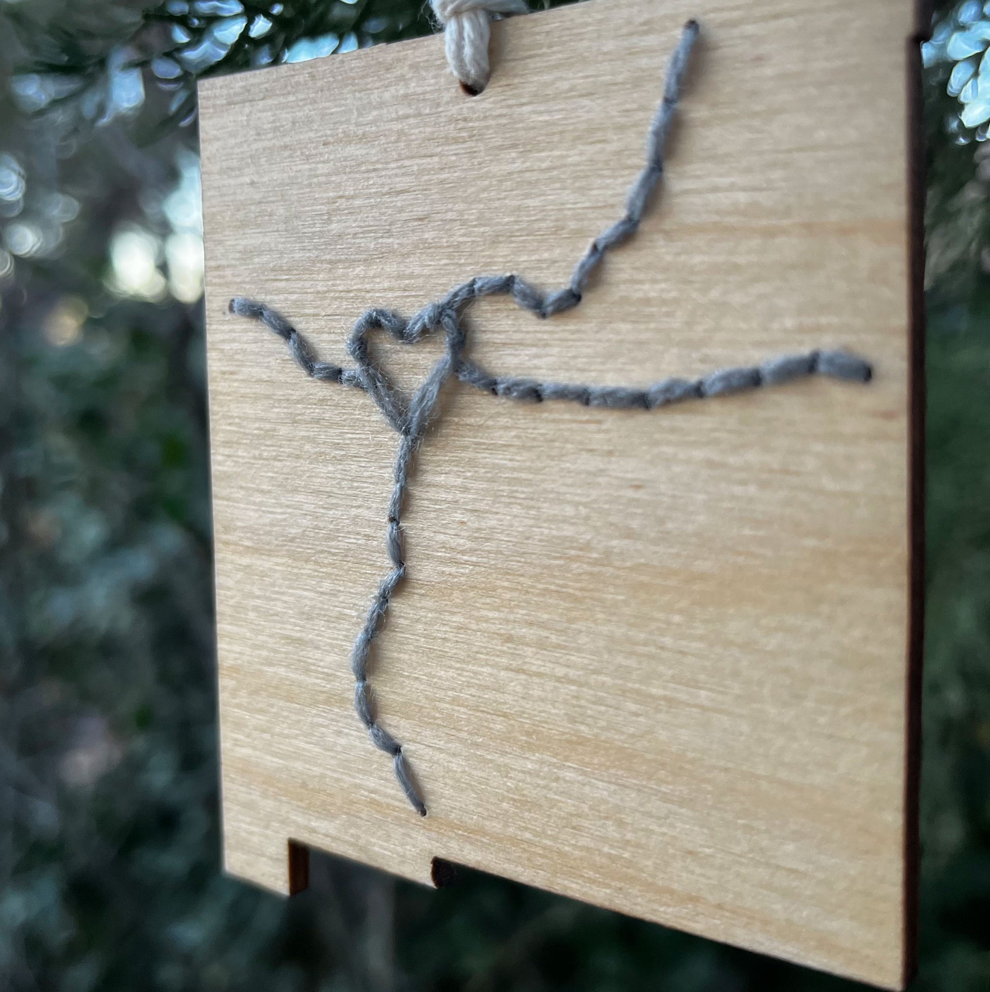 close up view of a laser cut wood ornament in the shape of New Mexico, hand embroidered in grey with a heart where Albuquerque is, and Interstate 25 and 40, with an off white cord loop hanging on a pine tree