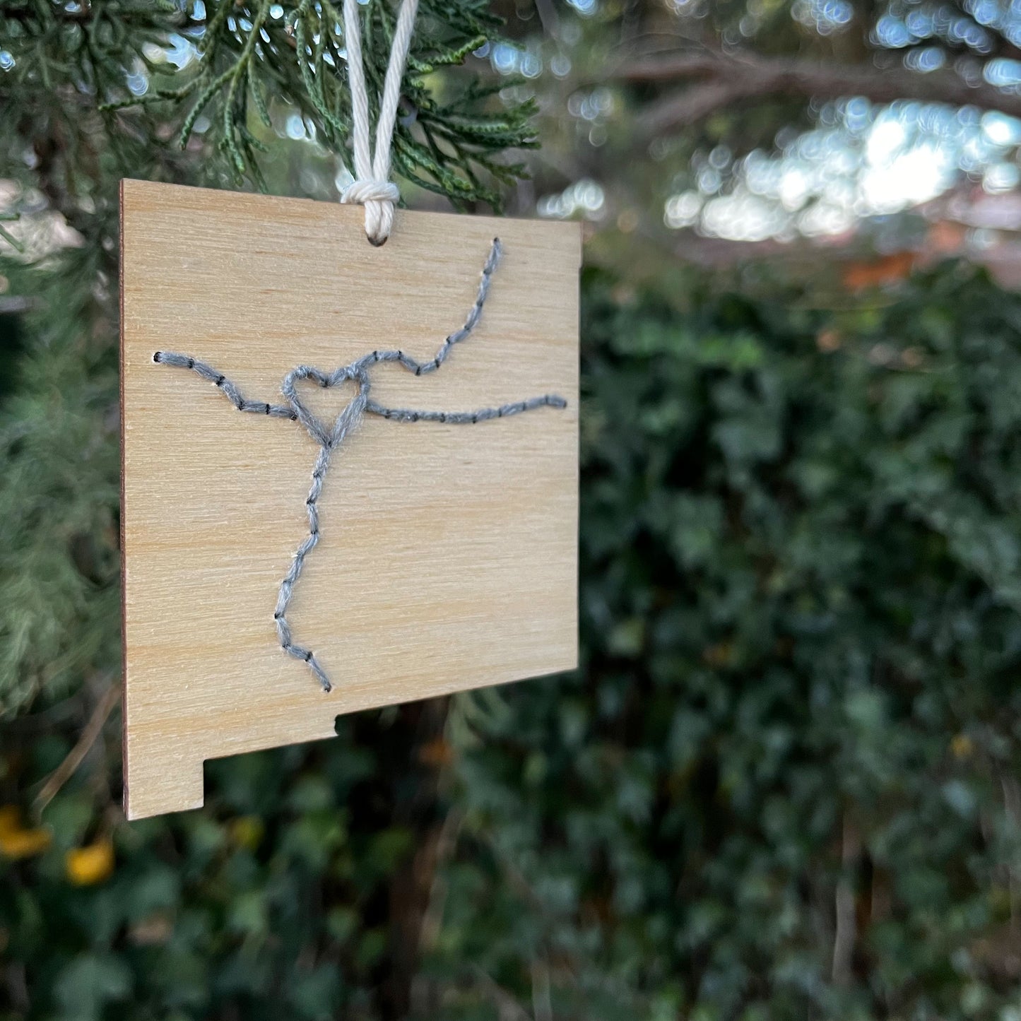 a laser cut wood ornament in the shape of New Mexico, hand embroidered in grey with a heart where Albuquerque is, and Interstate 25 and 40, with an off white cord loop hanging on a pine tree