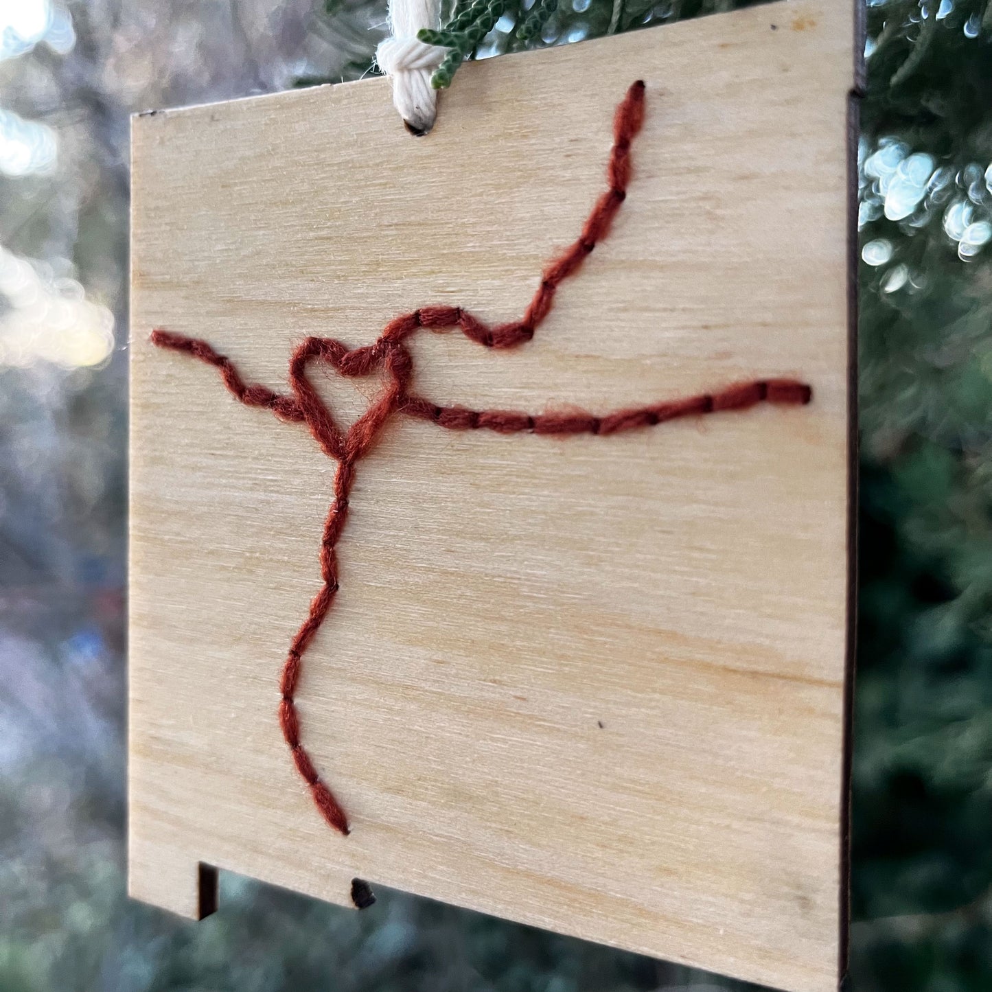 close up view of a laser cut wood ornament in the shape of New Mexico, hand embroidered in rust red with a heart where Albuquerque is, and Interstate 25 and 40, with an off white cord loop hanging on a pine tree