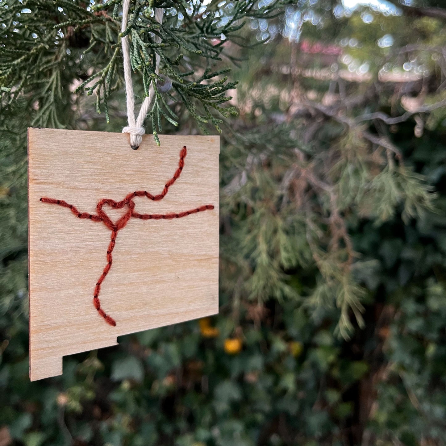 a laser cut wood ornament in the shape of New Mexico, hand embroidered in rust red with a heart where Albuquerque is, and Interstate 25 and 40, with an off white cord loop hanging on a pine tree