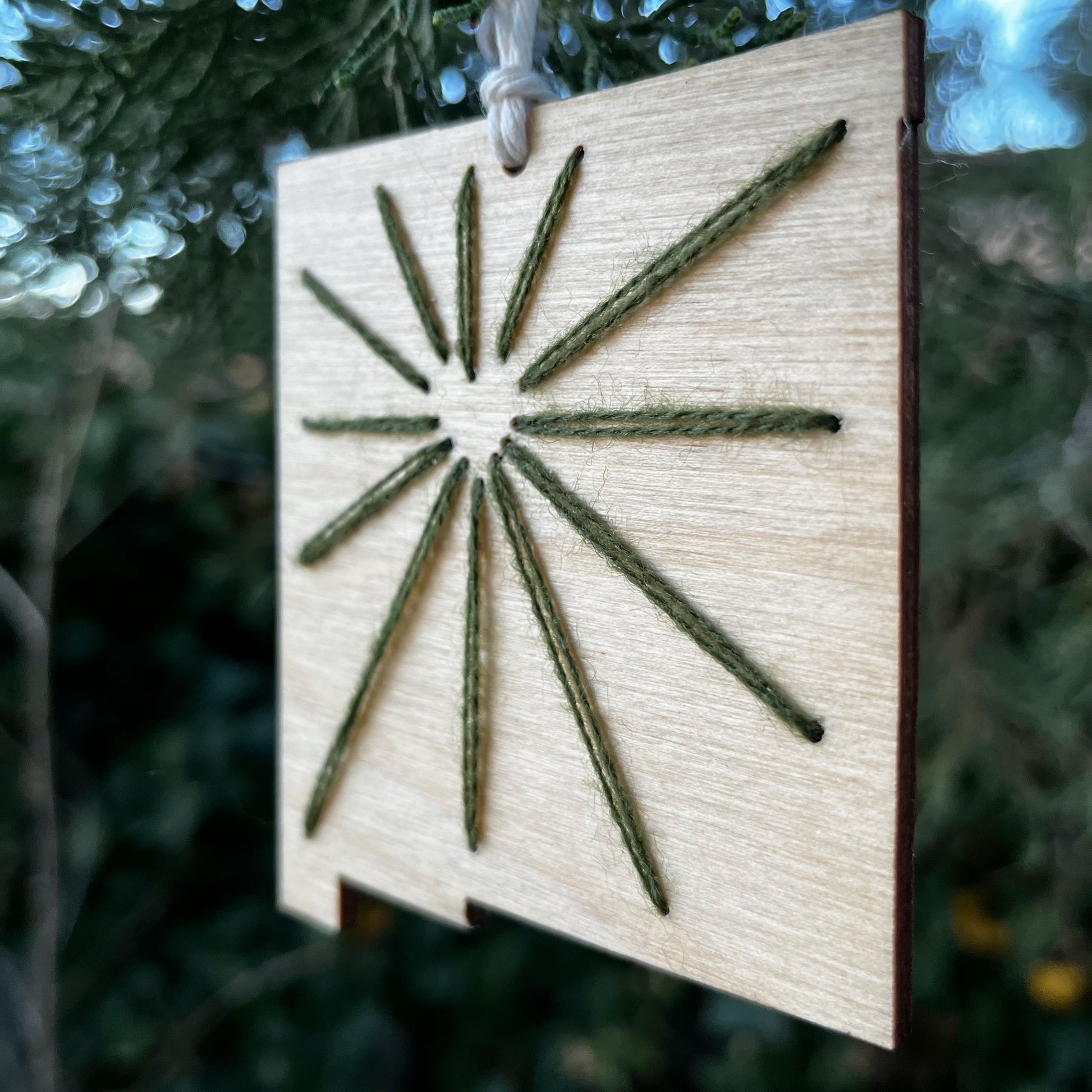 close up of a laser cut wood ornament in the shape of New Mexico, hand embroidered in green with negative space in the shape of a heart where Albuquerque is, and lines coming out of it like sunbeams, with an off white cord loop hanging on a pine tree
