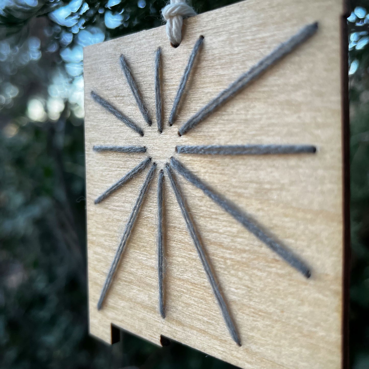 close up of a laser cut wood ornament in the shape of New Mexico, hand embroidered in grey with negative space in the shape of a heart where Albuquerque is, and lines coming out of it like sunbeams, with an off white cord loop hanging on a pine tree