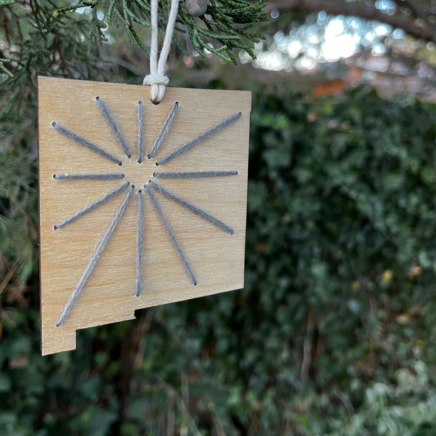 a laser cut wood ornament in the shape of New Mexico, hand embroidered in grey with negative space in the shape of a heart where Albuquerque is, and lines coming out from it like sunbeams, with an off white cord loop hanging on a pine tree