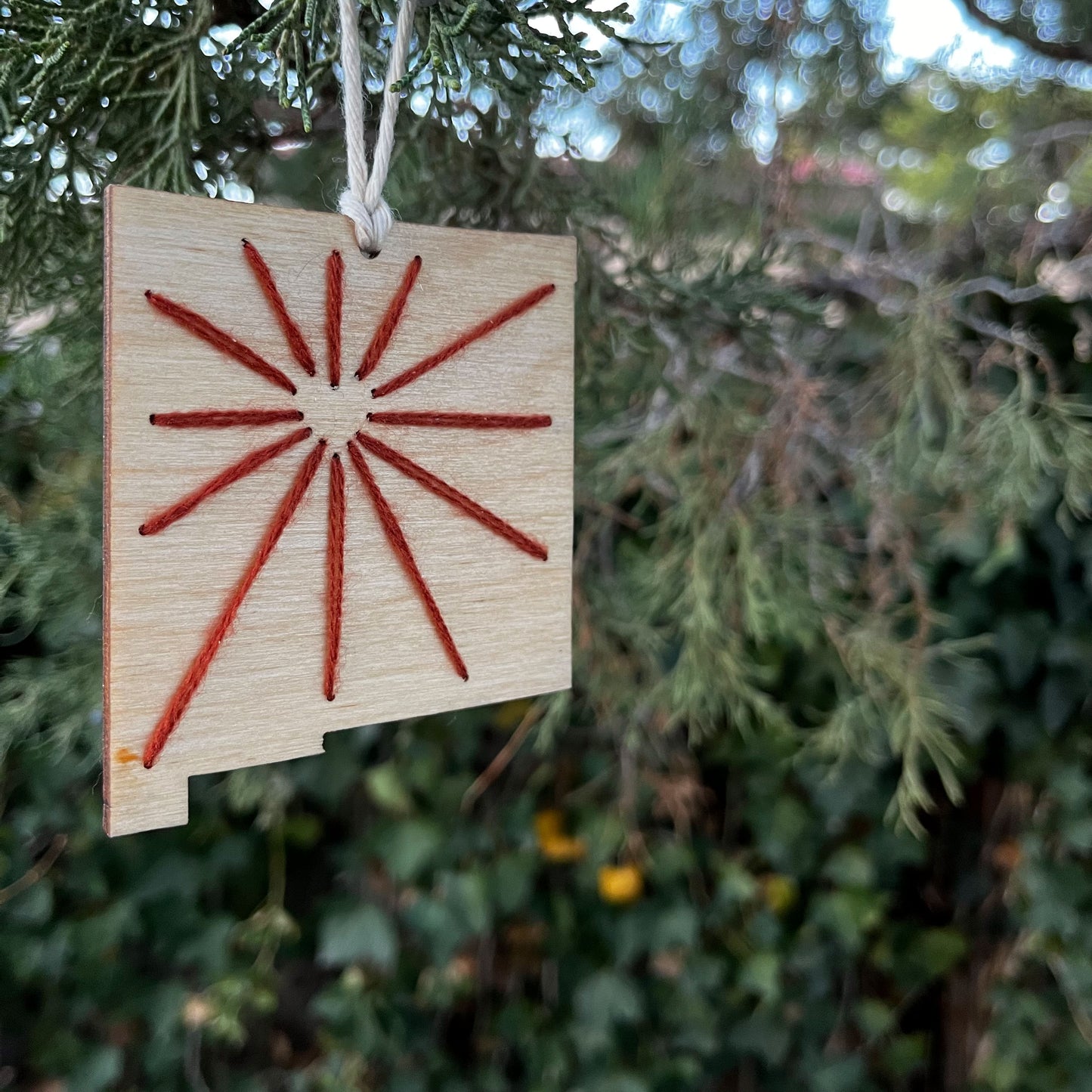 a laser cut wood ornament in the shape of New Mexico, hand embroidered in rust red with negative space in the shape of a heart where Albuquerque is, and lines coming out from it like sunbeams, with an off white cord loop hanging on a pine tree