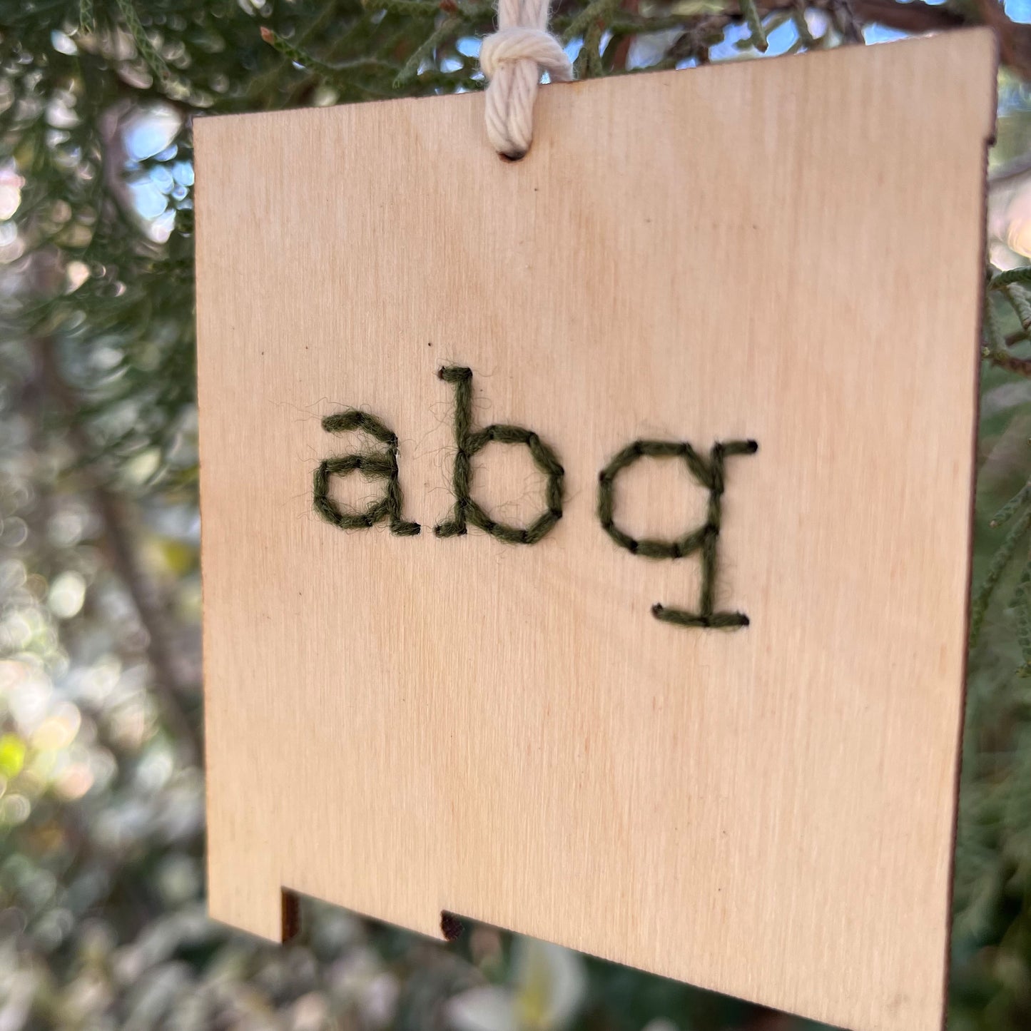 close up view of a laser cut wood ornament in the shape of New Mexico, hand embroidered in pine green with the letters abq for Albuquerque, with an off white cord loop hanging on a pine tree