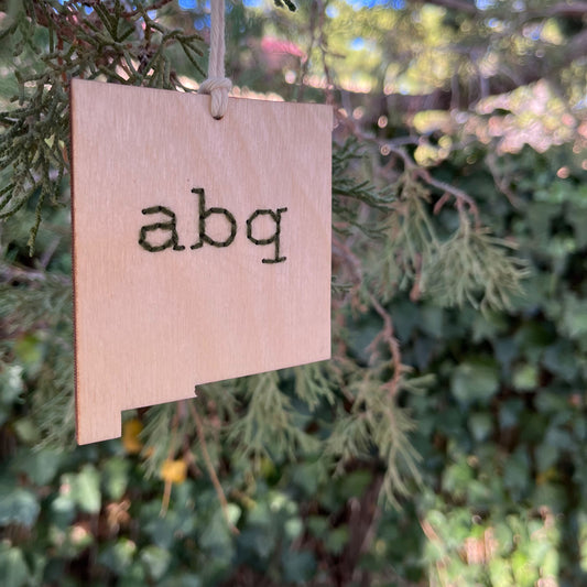 a laser cut wood ornament in the shape of New Mexico, hand embroidered in pine green with the letters abq for Albuquerque, with an off white cord loop hanging on a pine tree