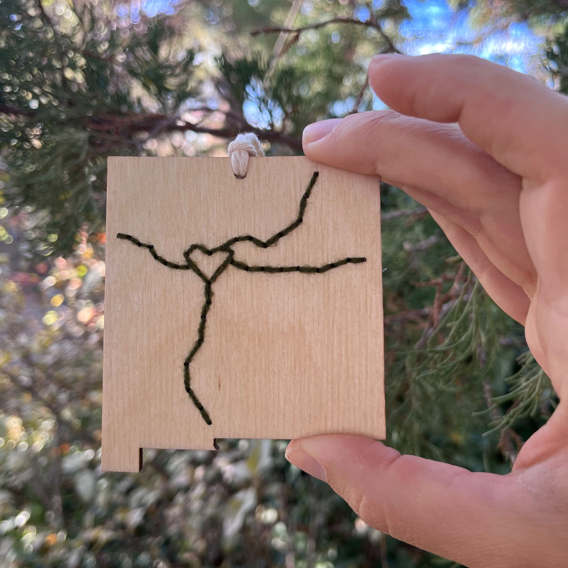 a hand holding a laser cut wood ornament in the shape of New Mexico, hand embroidered in pine green with a heart where Albuquerque is, and Interstate 25 and 40, with an off white cord loop, with a pine tree behind it