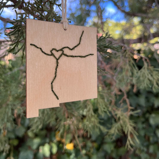 a laser cut wood ornament in the shape of New Mexico, hand embroidered in pine green with a heart where Albuquerque is, and Interstate 25 and 40, with an off white cord loop hanging on a pine tree