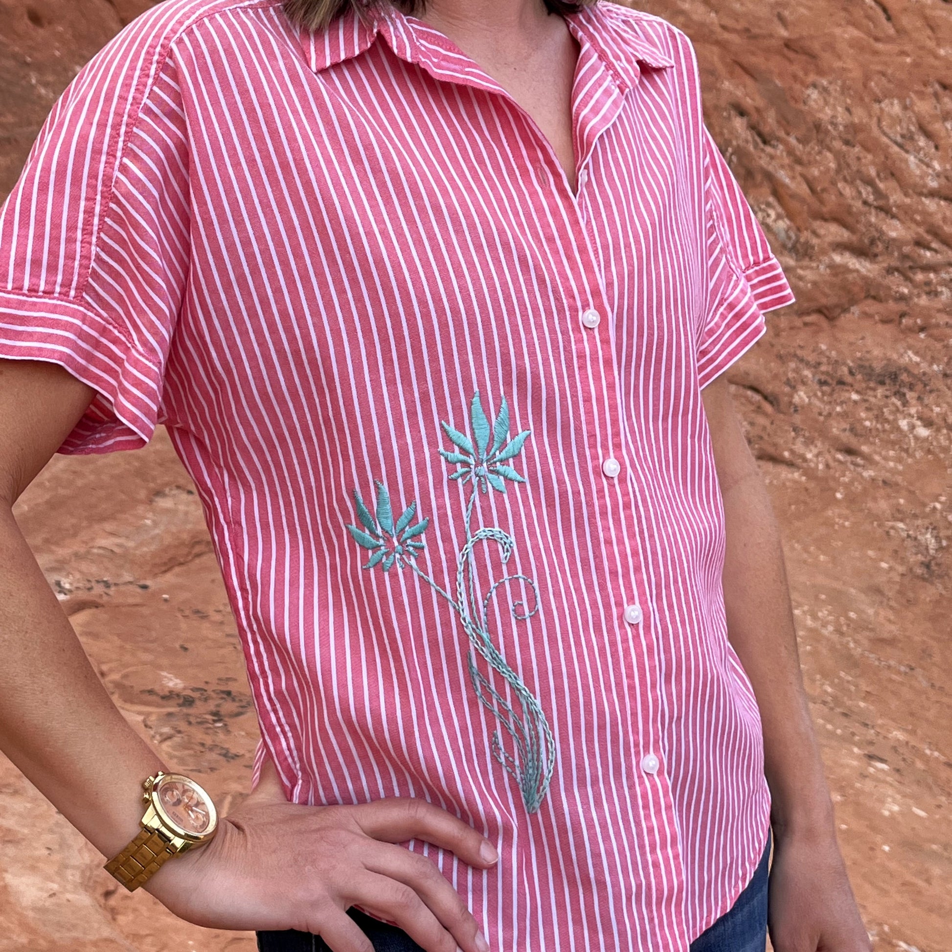 a woman's torso in a red and white striped button-up short sleeve shirt, with two hand embroidered long stemmed flowers in aqua on the lower front right side of the shirt, her right hand on her hip with a gold watch