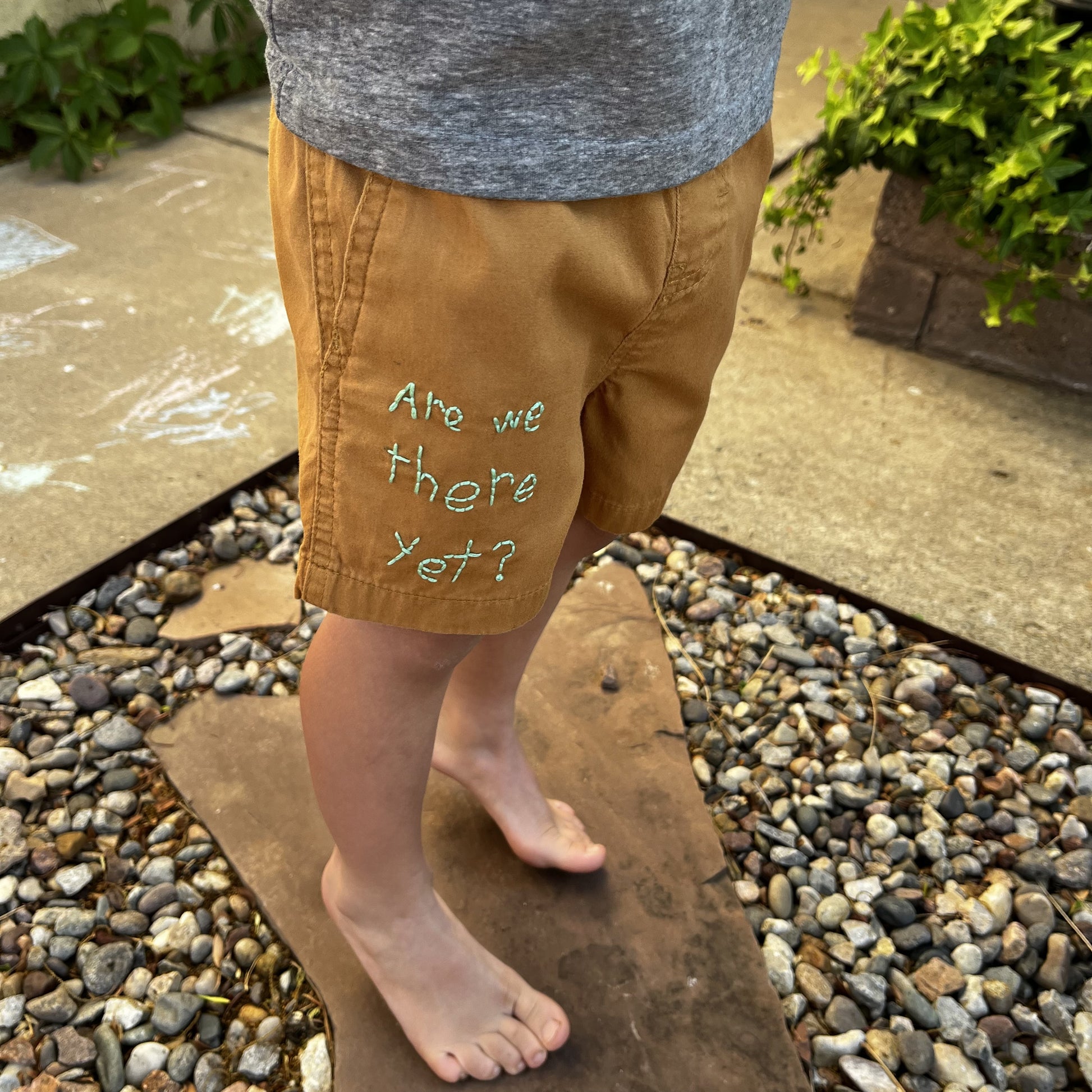 camel colored shorts on a toddler aged child, with the words "are we there yet?" hand embroidered on the lower right leg in green, the child is standing on a flagstone paver in rocks next to a sidewalk